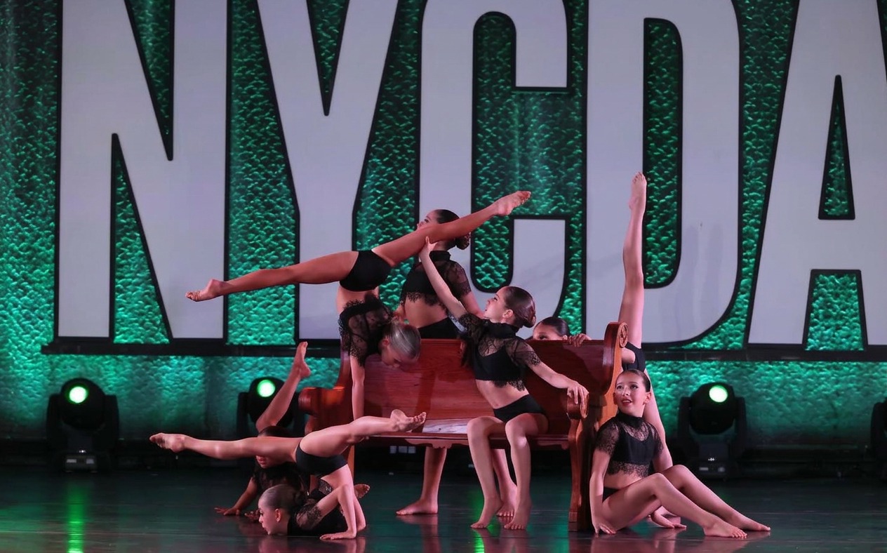 A group of dancers in black costumes performing a contemporary routine on a competition stage