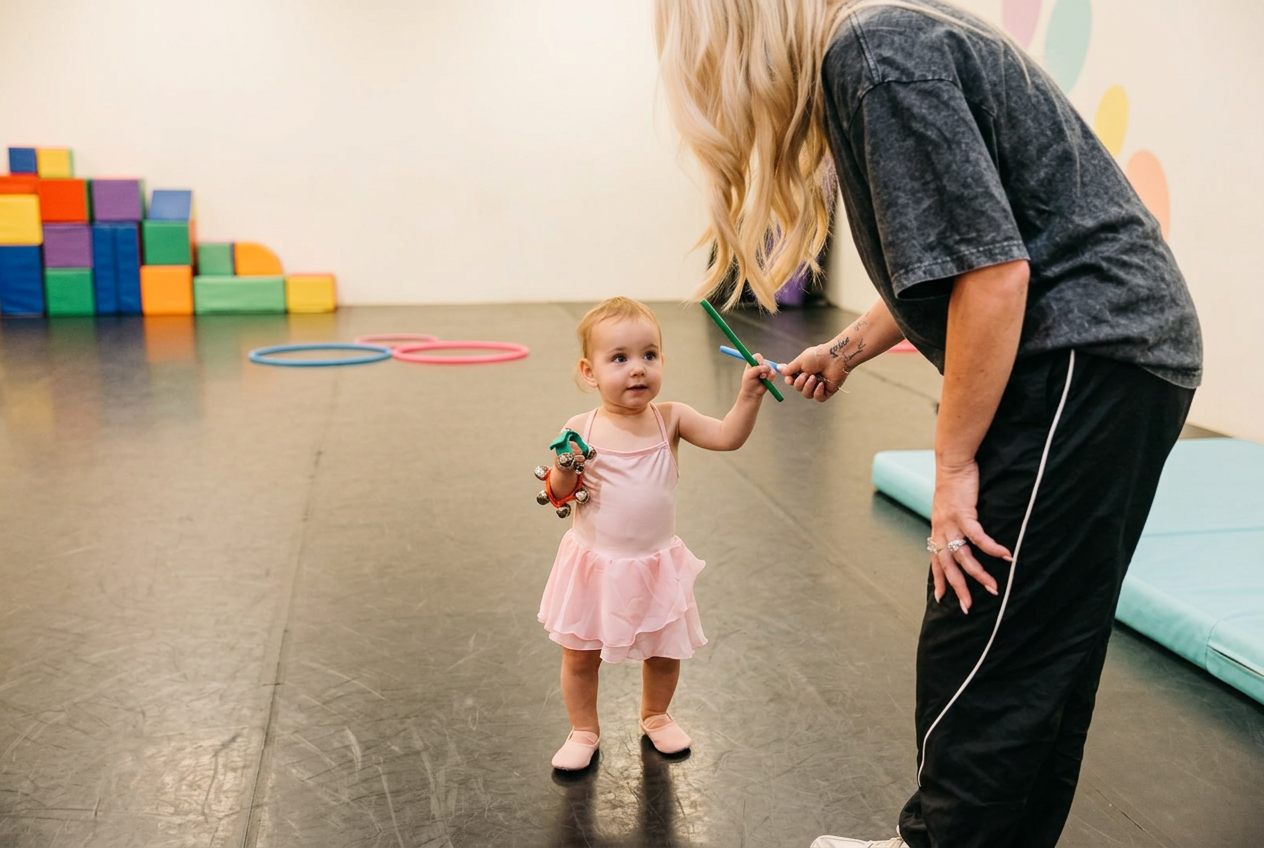 A young dancer holding bells while reaching toward a teacher in a bright dance studio